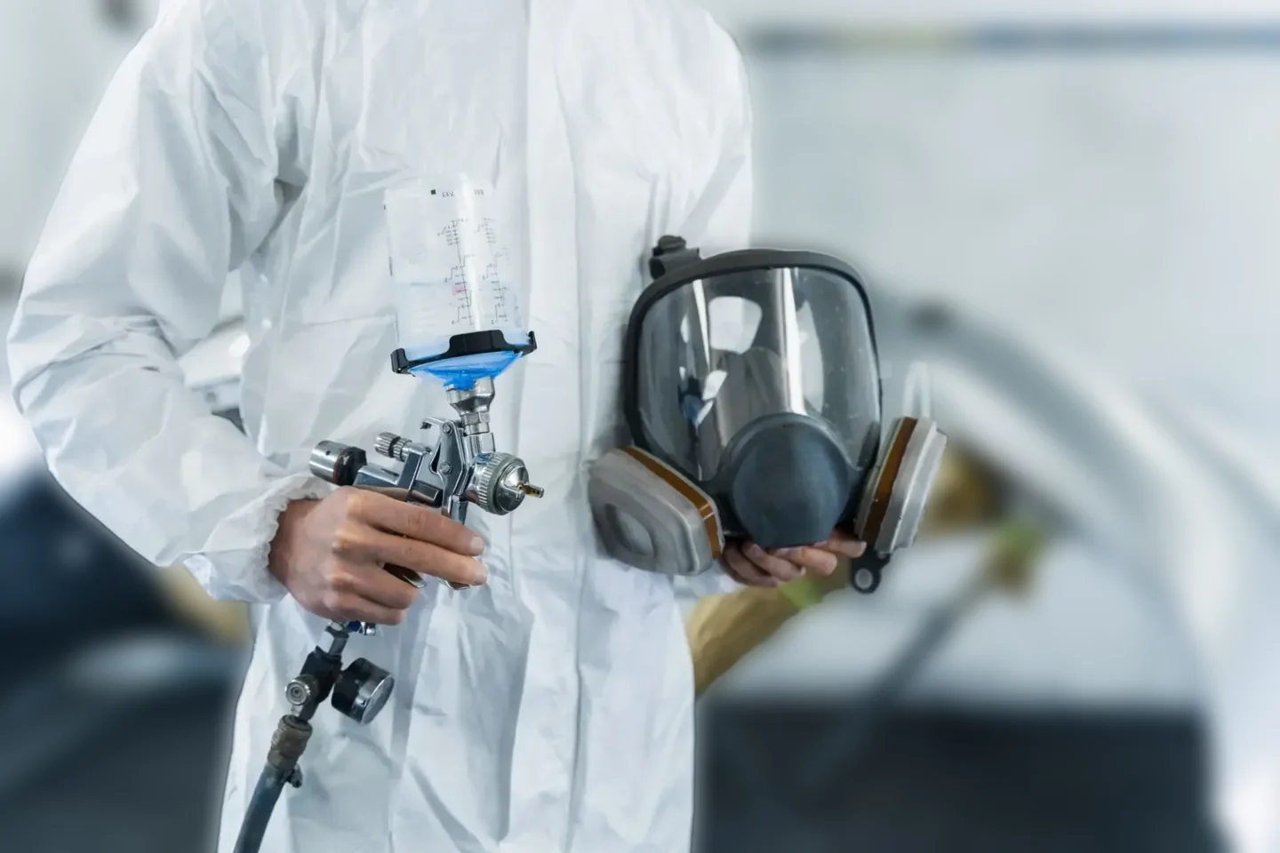 Person in protective suit holding a respirator mask and a spray gun in a laboratory or industrial setting.